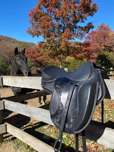 Freeform Sassenach treeless saddle in black sitting on a fence with a black horse and trees in the background.