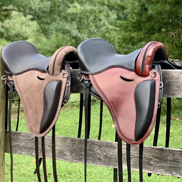 Two Freeform Treeless Saddles sitting on a fence in a green field, one brown and tan and one burgandy and black. Handmade in Italy.
